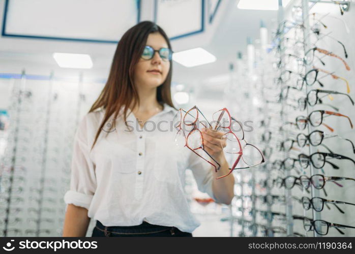 Young female buyer holds many glasses in hand, optics store, showcase with spectacles on background. Professional eyecare in glasses shop concept, eyeglasses choice. Young female buyer holds many glasses in hand