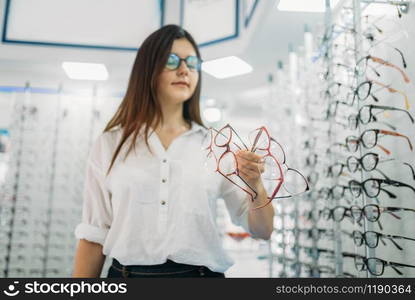 Young female buyer holds many glasses in hand, optics store, showcase with spectacles on background. Professional eyecare in glasses shop concept, eyeglasses choice. Young female buyer holds many glasses in hand