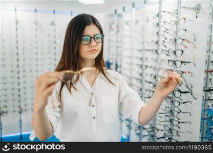 Young female buyer holds many glasses in hand, optics store, showcase with spectacles on background. Professional eyecare in glasses shop concept, eyeglasses choice. Young female buyer holds many glasses in hand