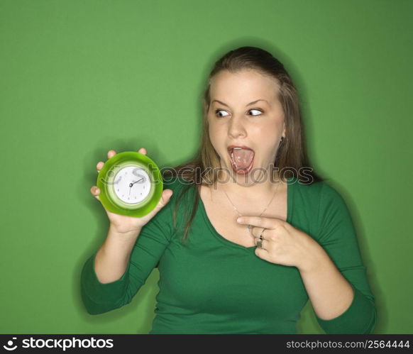 Young female Caucasian adult holding and pointing to clock.