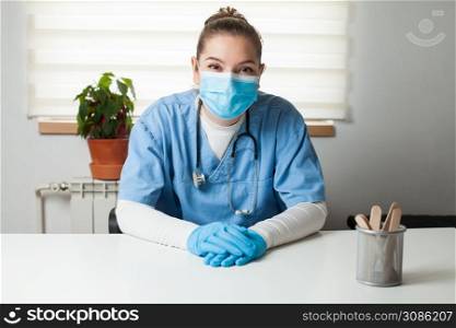 Young female caucasian General Practitioner sitting by her desk in office,wearing blue uniform,protective gloves & face mask,virtual tele visit via video call,GP doctor on demand telemedicine concept