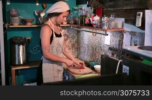 Young female chef cutting red fresh pepper on wooden cutting board in the kitchen