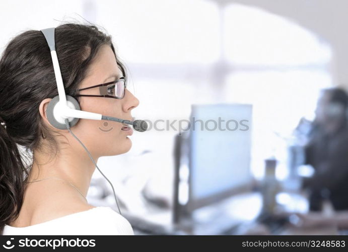 Young female customer service representative recieves calls on a headset while an IT specialist works on a computer in the background.