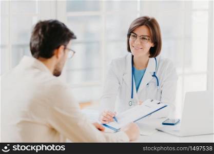 Young female doctor consults patient, shows information and prescription in clipboard, talk about medical checkup, pose at clinic, explains health insurance contract, read service agreement.