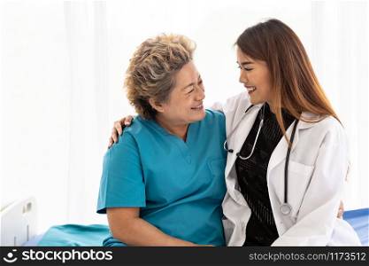Young female doctor in uniform hugging and smiling to old elderly woman patient during a visit in hospital ward
