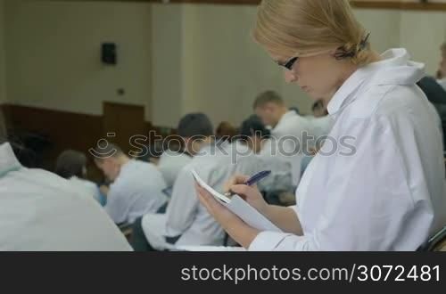 Young female doctor is listening the main speech of the medical conference and writing it down in notebook.