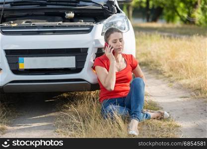 Young female driver sitting on ground next to broken car and calling for help