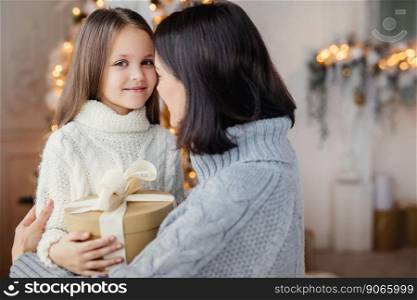 Young female embraces her little child, gives present, congratulates with New Year, proud to have such beautiful and clever daughter. Happy kid spends time with mother. Childhood and holidays