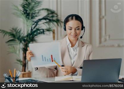 Young female employee economist in elegant suit and headset having online presentation, pointing at financial report with rising stats during video call on laptop and feeling sitisfied with results. Female economist having online presentation while working at office
