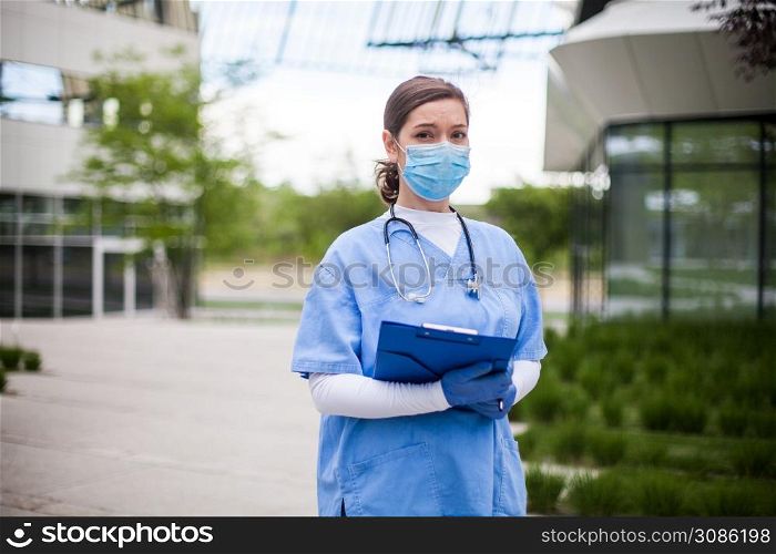 Young female EMS key worker doctor in front of healthcare ICU facility,wearing protective face mask holding medical patient lab health check form,COVID-19 pandemic outbreak crisis PPE shortage in UK