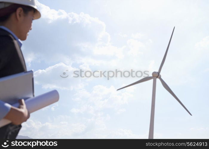 Young female engineer holding blueprints and checking wind turbines on site