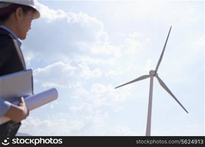 Young female engineer holding blueprints and checking wind turbines on site