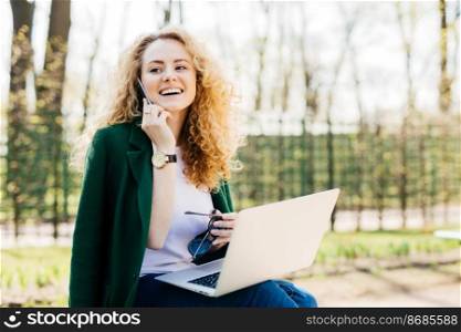 Young female entrepreneur with fluffy hair sitting outside in front of open generic laptop computer talking on mobile phone with happy look holding sunglasses in hand using laptop looking aside