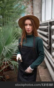 Young female florist in apron with shovel in hands against greenhouse on background. Female florist with shovel in hands