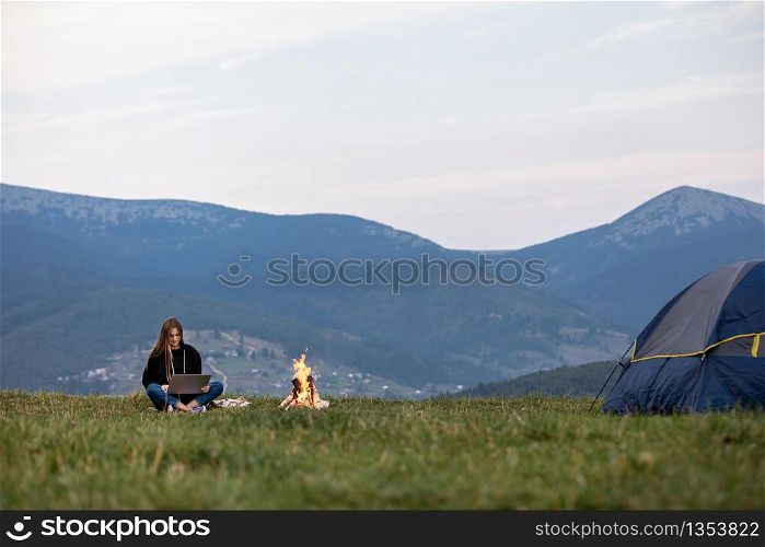 young female freelancer working on laptop in the mountains in the evening. Tourist girl sitting near campfire and having fun. Copy space. young female freelancer working on laptop in the mountains in the evening. Tourist girl sitting near campfire and having fun. Copy space.