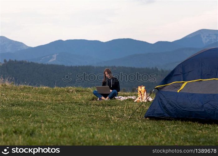 young female freelancer working on laptop in the mountains in the evening. Tourist girl sitting near campfire and tent. Copy space.