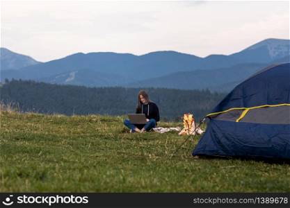 young female freelancer working on laptop in the mountains in the evening. Tourist girl sitting near campfire and tent. Copy space.