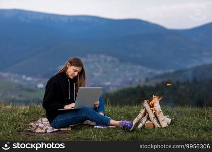young female freelancer working on laptop in the mountains in the evening. Tourist girl sitting near c&fire and having fun. Copy space. young female freelancer working on laptop in the mountains in the evening. Tourist girl sitting near c&fire and having fun. Copy space.