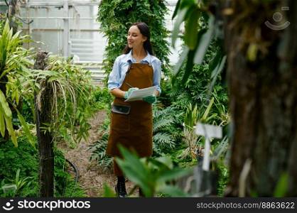 Young female gardener working in greenhouse growing tropical plants. Woman making notes in notebook during research experiment. Gardener working in greenhouse growing tropical plants