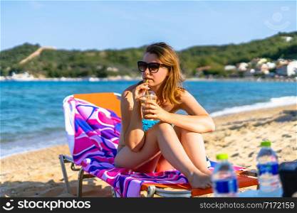 Young female girl woman hand holding a frappe or ice coffee at the beach in swimsuit bikini while relaxing in a sunbed at vacation in sunny day drinking by the sea