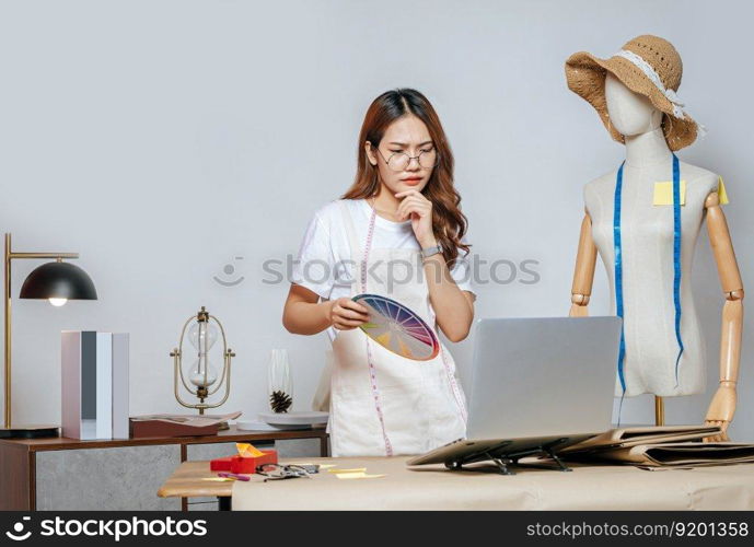 young female in eyeglasses fashion designer or tailor working with laptop computer to search idea or online orders, full of tailoring tools with mannequin and equipment on the desk in studio