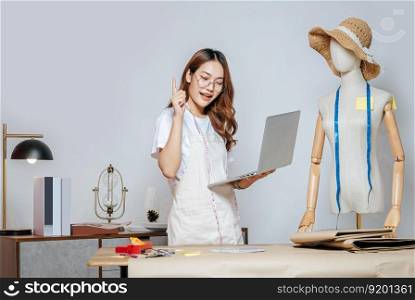 young female in eyeglasses fashion designer or tailor working with laptop computer to search idea or online orders, full of tailoring tools with mannequin and equipment on the desk in studio