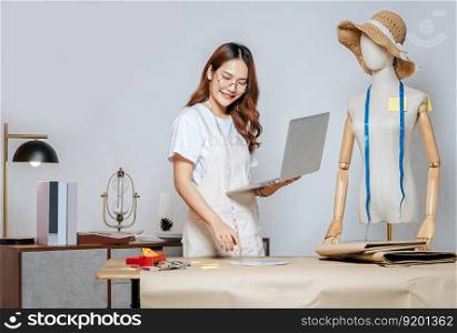 young female in eyeglasses fashion designer or tailor working with laptop computer to search idea or online orders, full of tailoring tools with mannequin and equipment on the desk in studio