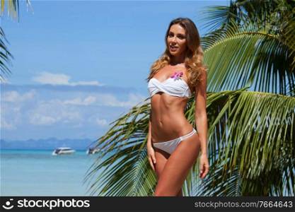 Young female in yellow bikini enjoying sunny day under palm tree on the tropical beach. Girl in bikini under palm tree
