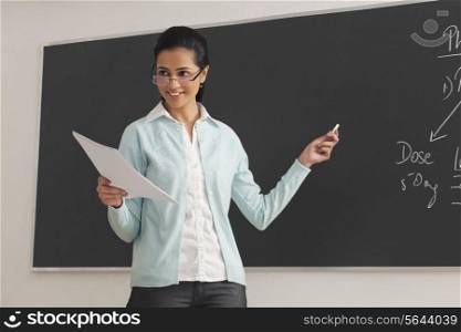 Young female lecturer holding papers and chalk against blackboard