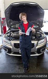Young female mechanic reading paper with open bonnet of car in garage