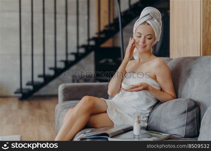 Young female model applies face cream, has perfect body, healthy smooth skin, sits in cozy room on sofa, wrapped in towel, reads magazine, takes care of complexion. Cosmetology and beauty concept