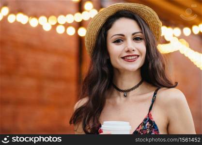 Young female model with dark long hair, hazel eyes and red lips, wearing necklace, summer hat and dress having delightful expression while posing at outdoor cafe. People, rest, emotions concept