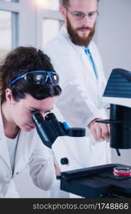 Young female scientist doing test in laboratory, male scientist taking notes near female colleague working with microscope