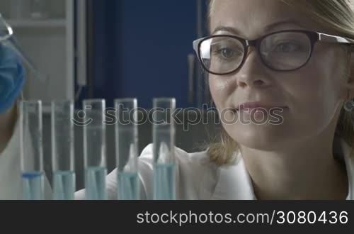 Young female scientist in protective glasses and gloves dropping liquid substance into test tubes with pipette in scientific chemical laboratory. Beautiful female scientific researcher observing the blue indicator color shift in glass tubes in lab