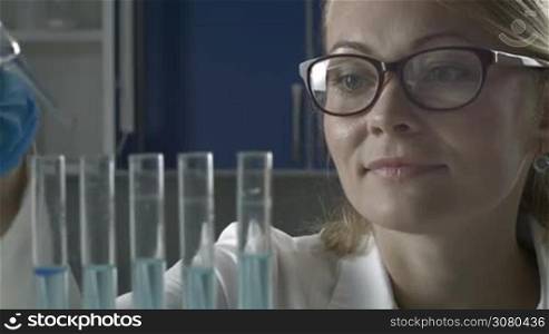Young female scientist in protective glasses and gloves dropping liquid substance into test tubes with pipette in scientific chemical laboratory. Beautiful female scientific researcher observing the blue indicator color shift in glass tubes in lab