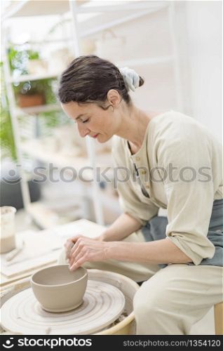 Young female sitting by table and making clay or ceramic mug in her working studio