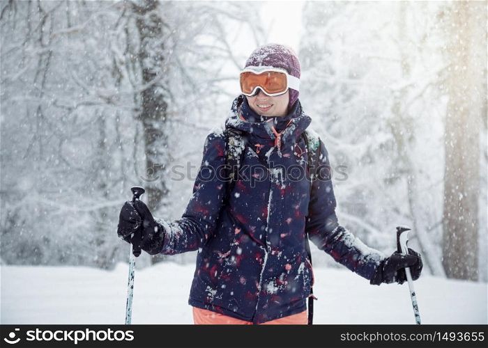 Young female skier standing on ski slope under snowfall, holding ski poles and smiling, close up