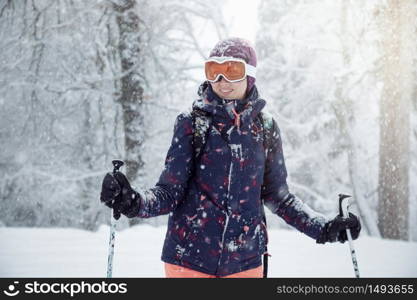 Young female skier standing on ski slope under snowfall, holding ski poles and smiling, close up