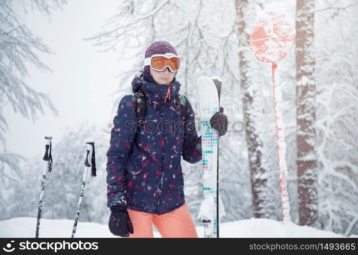 Young female skier standing on ski slope under snowfall, holding ski poles and smiling, close up