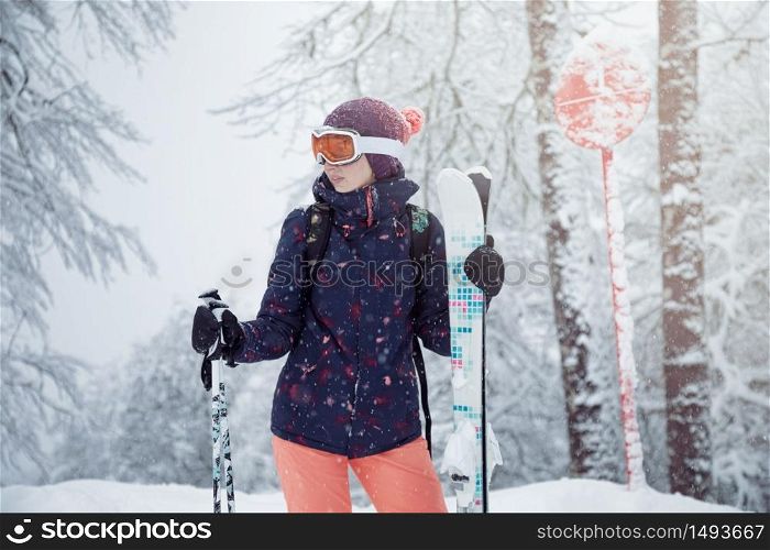 Young female skier standing on ski slope under snowfall, holding ski poles and smiling, close up