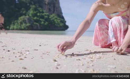 Young Female squatting down playing with sand picking up clam shell on white sand beach on sunny day. Young pretty girl playing with sea sand water waves. summer travel destinations. Low camera angle
