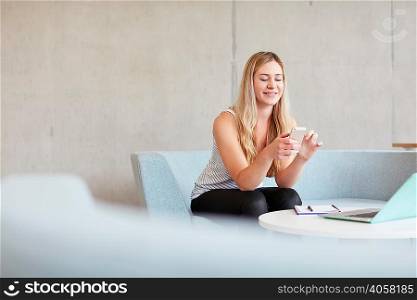 Young female student sitting on study space sofa reading smartphone texts at higher education college