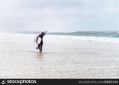 Young female surfer wearing wetsuit, holding surfboard under his arm, walking on beach after morning surfing session.