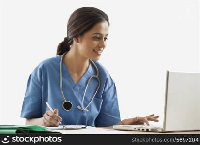Young female surgeon using laptop while writing notes at desk
