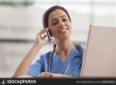 Young female surgeon using mobile phone while looking at laptop screen
