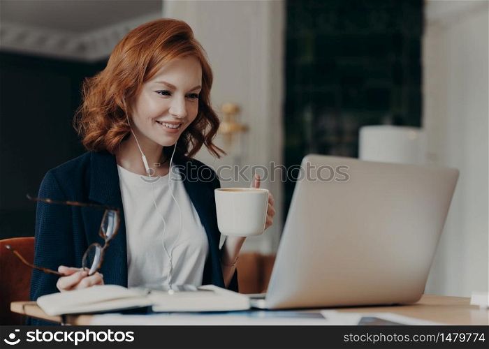 Young female teacher conducts lesson online, talks wih pupils via laptop computer, organizes video conference, teaches students, drinks aromatic coffee, holds glasses in hand, dressed formally