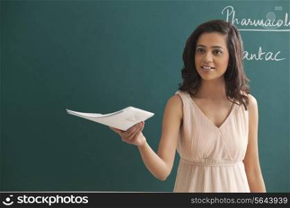 Young female teacher holding file while looking away against green board