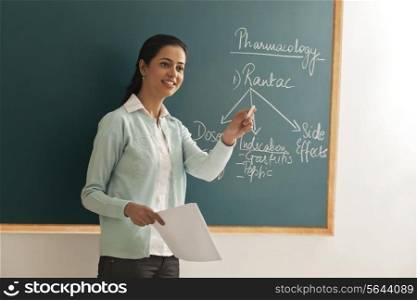 Young female teacher holding paper and chalk against green board
