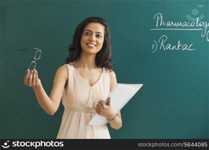 Young female teacher looking away while holding glasses and file against green board