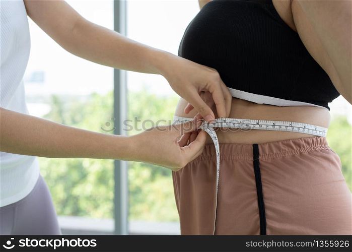 Young female trainer measuring fat layer of overweight woman with waistline at fitness